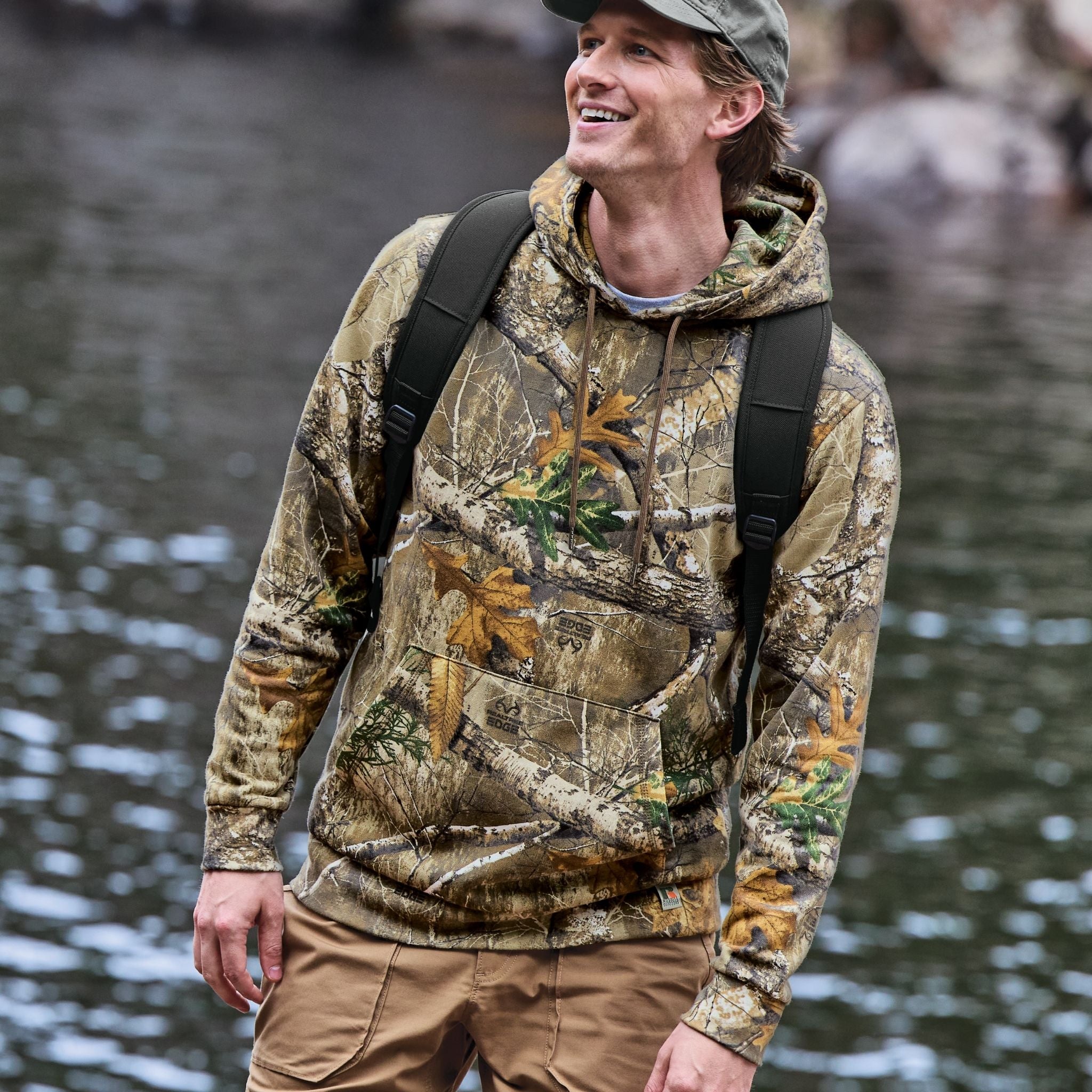 Man wearing a Realtree Edge camouflage hoodie and backpack standing by a body of water.